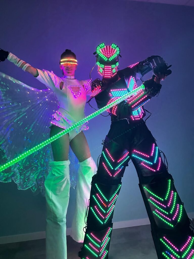Robot LED en zancos con luces multicolor y sable láser junto a bailarina con alas de luces LED y traje blanco